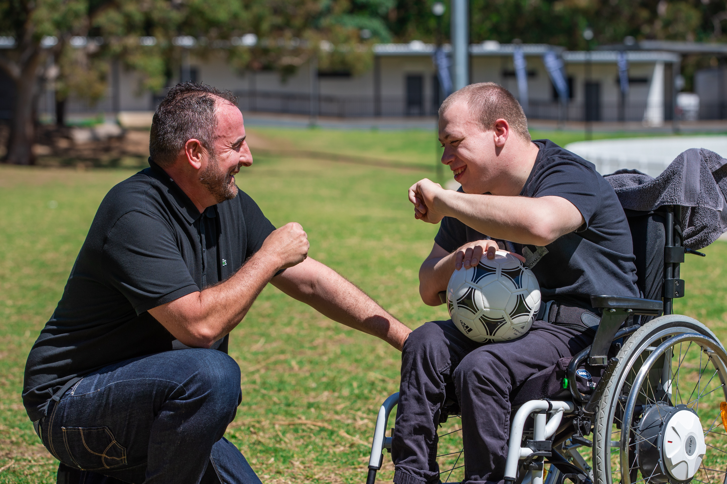 Corey and Mark are in the middle of a fist pump. They are at the soccer oval. Corey, seated in his wheelchair is holding a soccer ball on his lap.