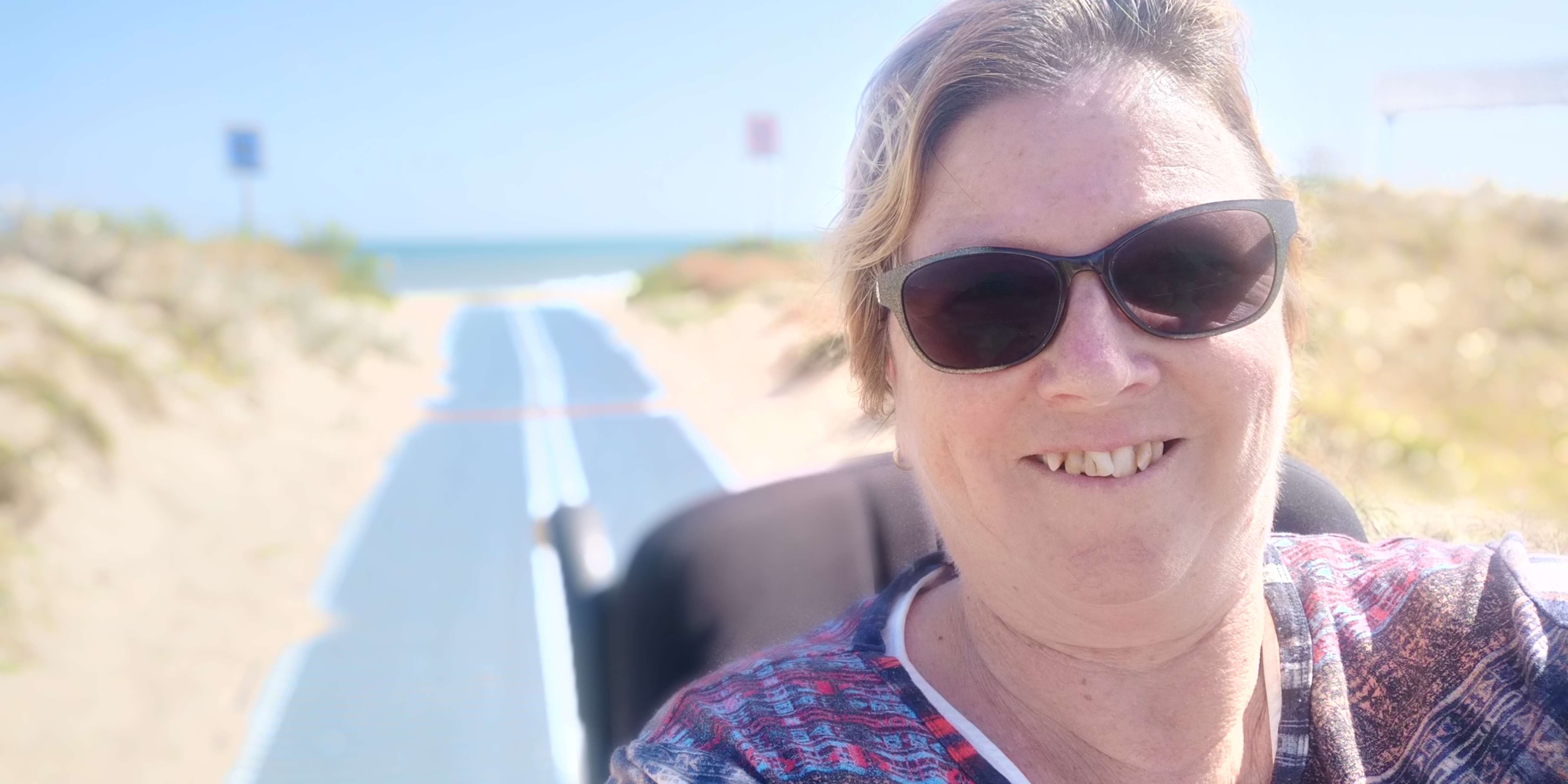 Lynne has light brown hair and is wearing sunglasses. She is smiling widely at the camera for this close up photo. Behind her, is an accessible beach mat that is stretched across the sand to the sea. 