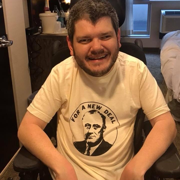 Todd has short hair and a beard. He's wearing a t-shirt with an image of American President Franklin D Roosevelt. The words 'For a new deal' surround the image. Todd is a wheelchair user. 
