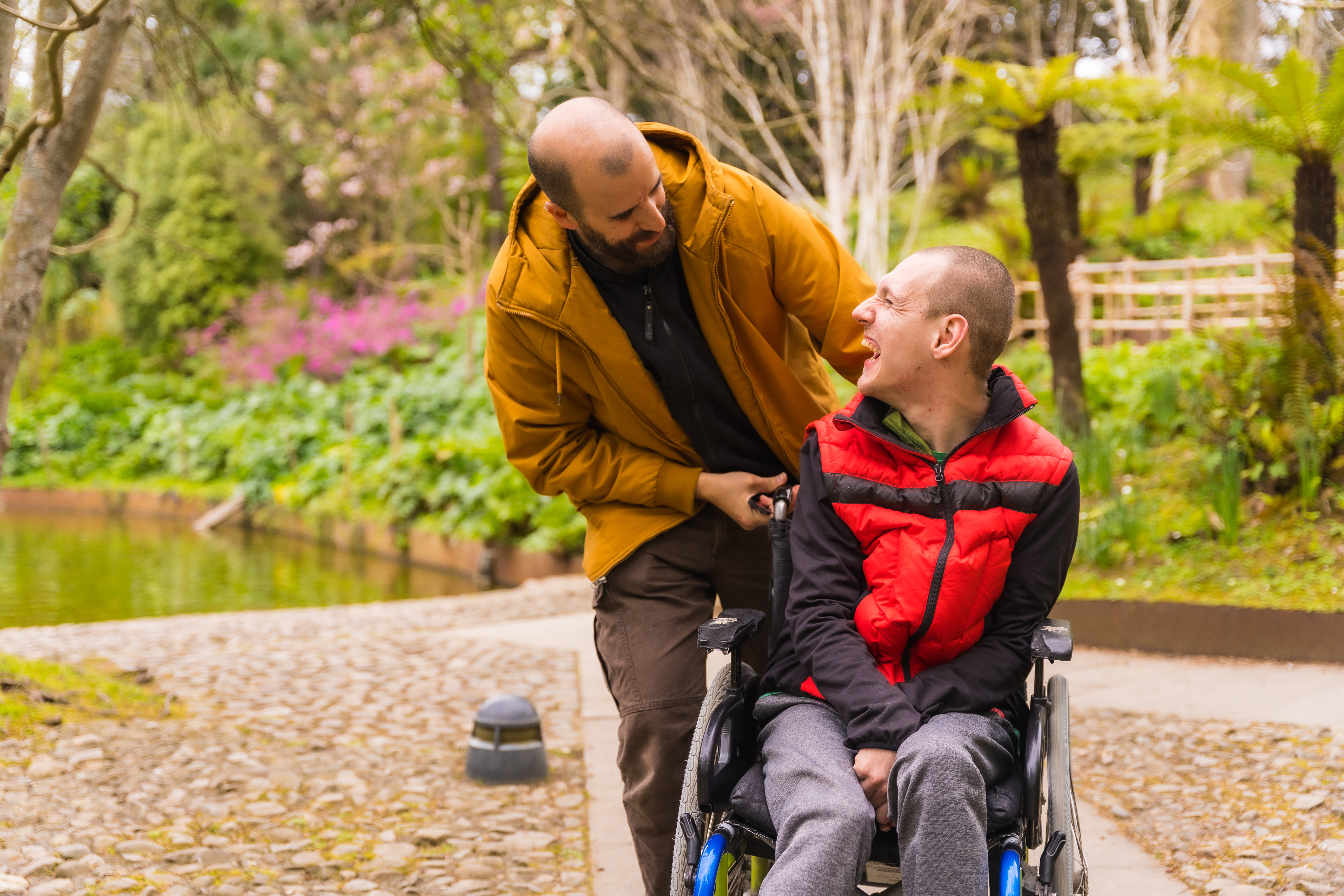 Two men with shaved short brown hair are smiling at each other. One is wearing a bright red puffer vest, black long sleeved shirt, grey tracksuit pants and is a wheelchair user. The other man is behind the first wearing a mustard yellow rain jacket. There are green trees and a park pond in the background.