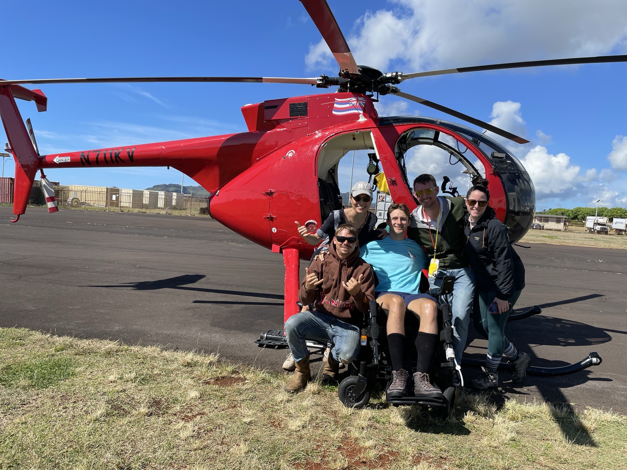 A group of friends, including wheelchair user Steve Ralph, pose in front of a red helicopter. 