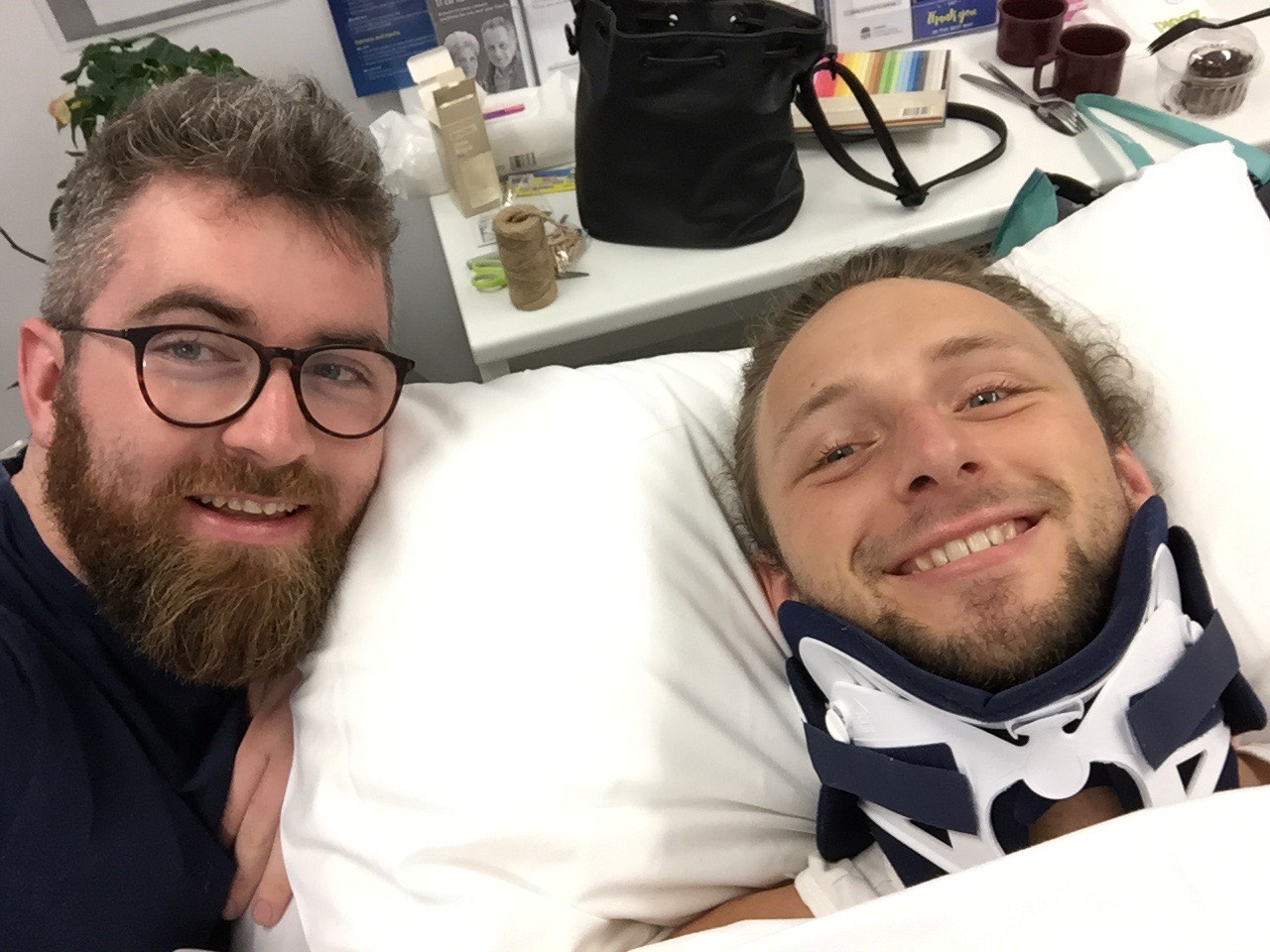 Steven is laying in bed and is wearing a neck brace. His friend, who has a beard and wears glasses, is taking a selfie photo with Steven. They are both smiling.