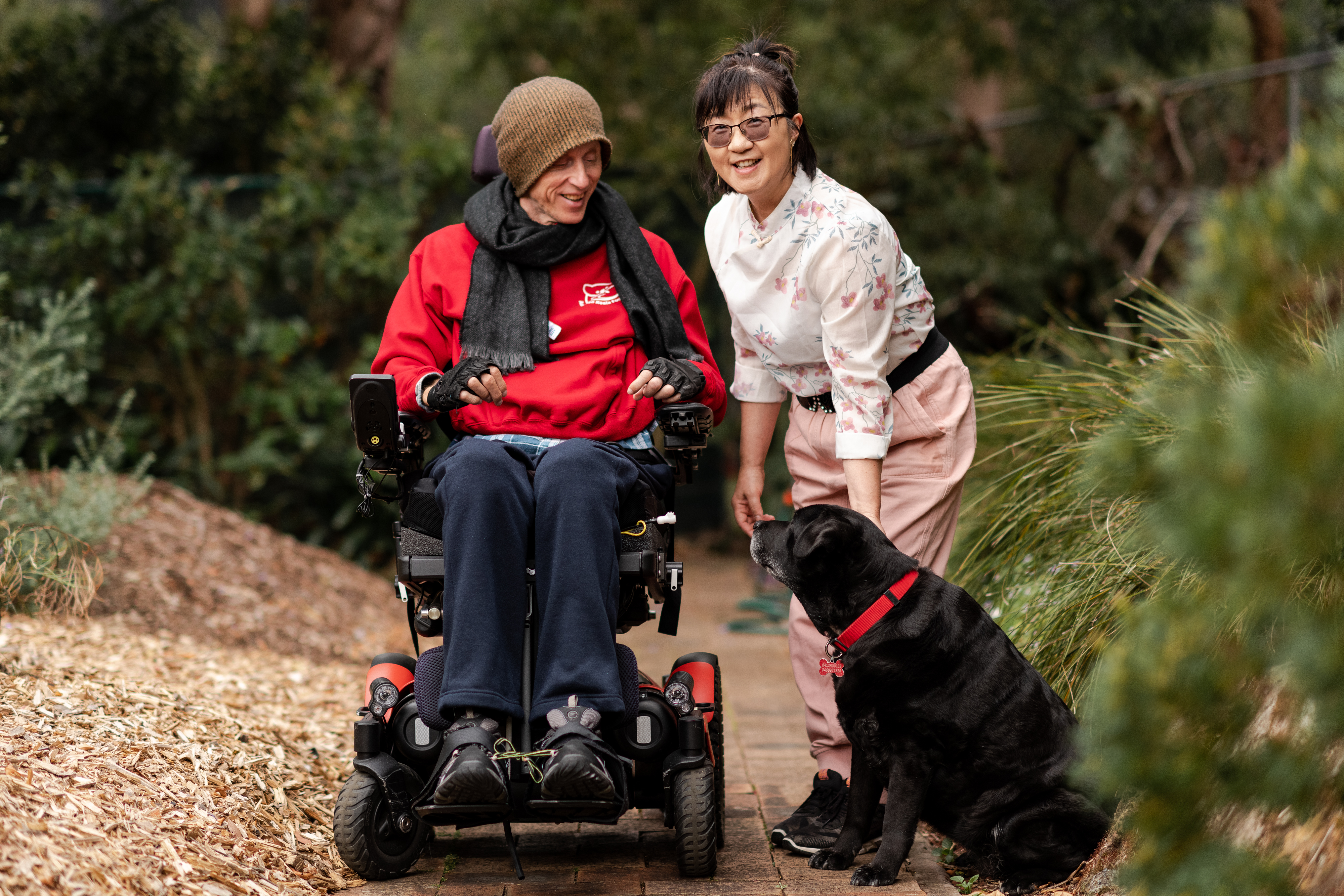 Annie and Stew are smiling together on a footpath with Stew's black labrador. Annie has short black hair, wears glasses and is wearing a white top with pink flowers and pink shorts. Stew is wearing a brown beanie, black scarf, red jumper and navy track pants. Stew is a wheelchair user.