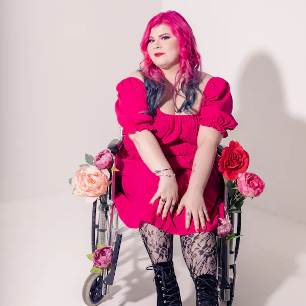 Zoe Simmons, a disability advocate and writer, is wearing a bright pink dress, floral fishnet stockings, and black heeled ankle boots. She has pale skin, long pink and blue hair, and bright makeup. She’s sitting in a silver and black manual wheelchair, adorned in fake flowers.