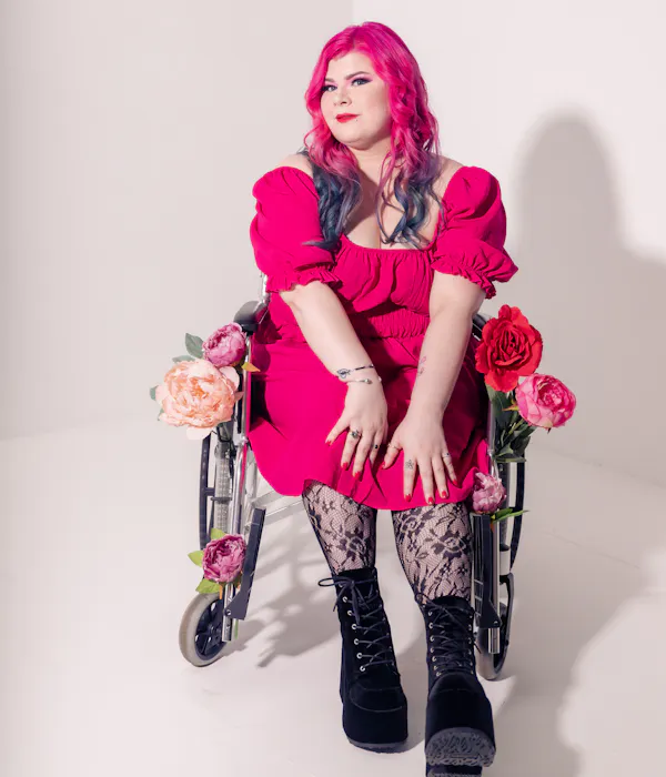 Zoe Simmons, a disability advocate and writer, is wearing a bright pink dress, floral fishnet stockings, and black heeled ankle boots. She has pale skin, long pink and blue hair, and bright makeup. She’s sitting in a silver and black manual wheelchair, adorned in fake flowers.