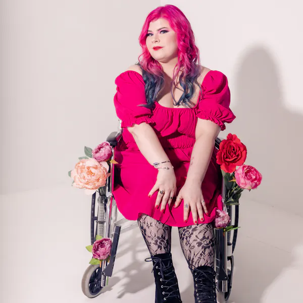 Zoe Simmons, a disability advocate and writer, is wearing a bright pink dress, floral fishnet stockings, and black heeled ankle boots. She has pale skin, long pink and blue hair, and bright makeup. She’s sitting in a silver and black manual wheelchair, adorned in fake flowers.