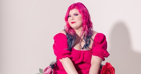 Zoe Simmons, a disability advocate and writer, is wearing a bright pink dress, floral fishnet stockings, and black heeled ankle boots. She has pale skin, long pink and blue hair, and bright makeup. She’s sitting in a silver and black manual wheelchair, adorned in fake flowers.