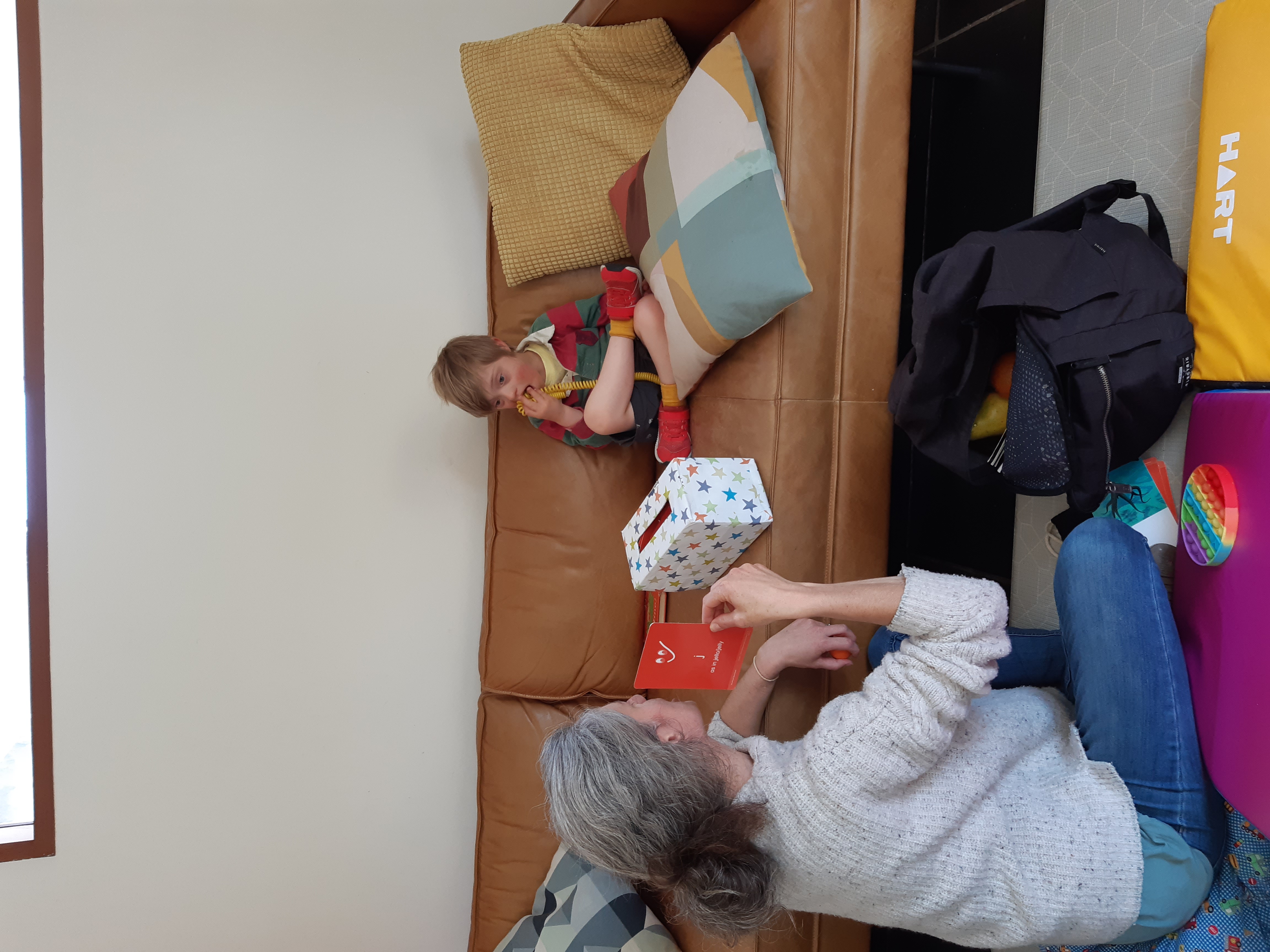 Oscar is sitting crossed legged on a couch. He is wearing a red and green striped shirt. Naomi is sitting in front of the couch on the floor facing Oscar. She is holding a red alphabet flash card and is wearing a grey woolen jumper and blue denim jeans. 