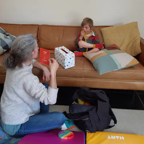 Oscar is sitting crossed legged on a couch. He is wearing a red and green striped shirt. Naomi is sitting in front of the couch on the floor facing Oscar. She is holding a red alphabet flash card and is wearing a grey woolen jumper and blue denim jeans.