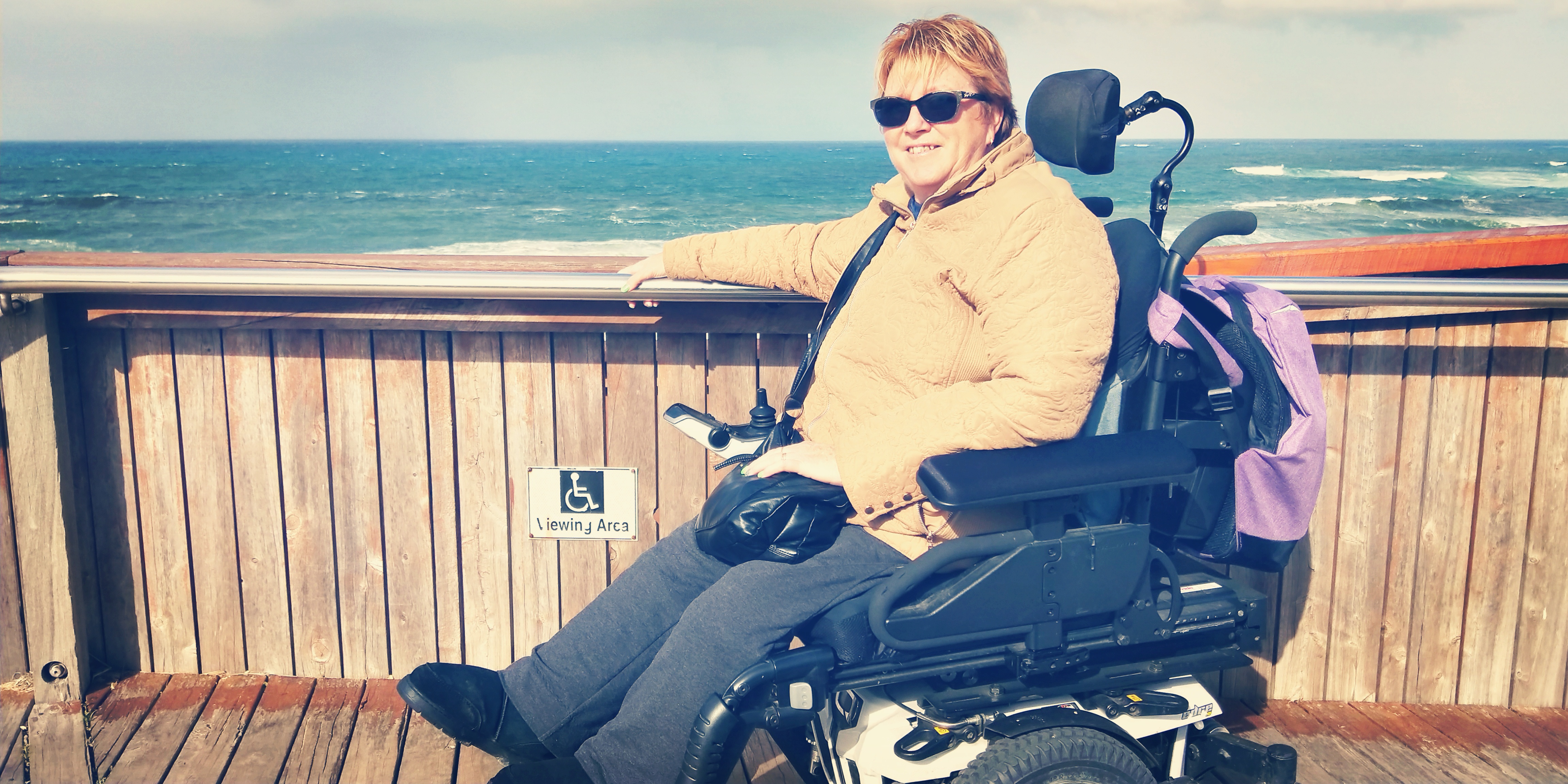 Lynne is wearing sunglasses and is rugged up in a caramel winter jacket and ugg boots. She is on a vantage point platform. Lynne is a wheelchair user. In the background is the ocean.  