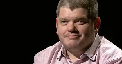 Disability activist Todd Winther smiles while on a television set, behind him is a black screen. He wears a pale red button-up shirt and a lapel microphone.