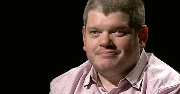 Disability activist Todd Winther smiles while on a television set, behind him is a black screen. He wears a pale red button-up shirt and a lapel microphone.