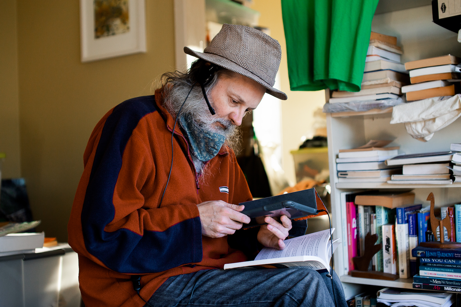 Hireup user Andrew is using an ipad with headphones. He also has a book open on his lap and is surrounded in the room by bookcases.