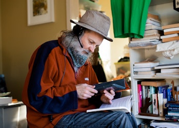 Hireup user Andrew is using an ipad with headphones. He also has a book open on his lap and is surrounded in the room by bookcases.