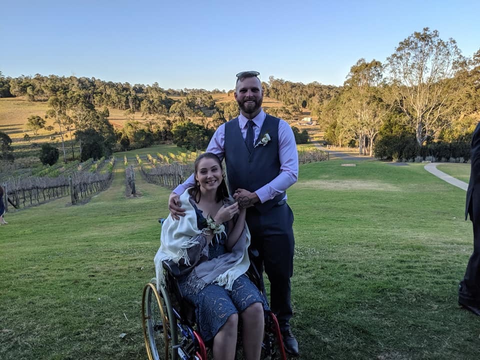 Megan wears a lace dress and shawl. A man wearing a tuxedo stands next to Megan and has his arms wrapped around her shoulders. They're at a vineyard at sunset. Megan is a wheelchair user. 