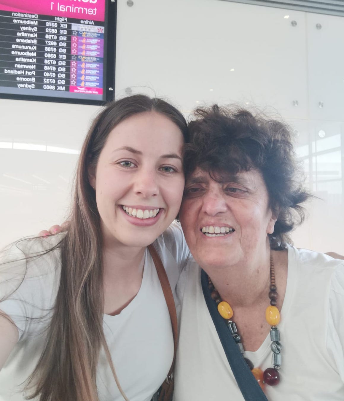 Tabitha and Gisele are embracing each other and smiling. Both are wearing white t-shirts. They are pictured in the Perth Airport.  