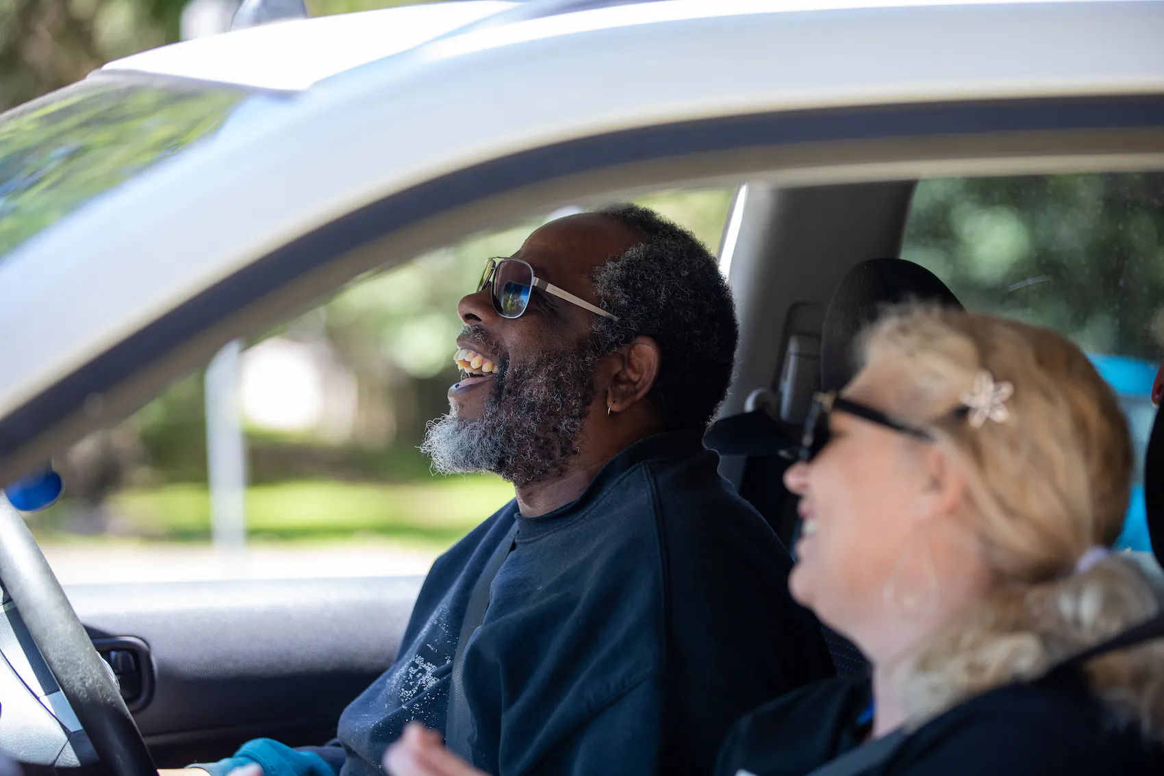 Caroline and Pat are in the front seat of the car. They are singing along and laughing to the music.