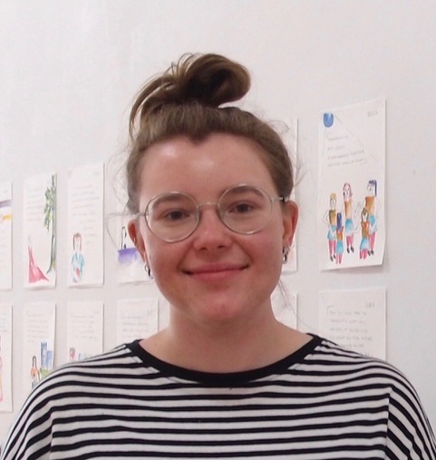Amelia Saward wears her long brown hair high on her head. She has glasses on and wears a striped black and white t-shirt. She smiles at the camera while standing in front of a wall with pages of drawing and writing stuck to it.  