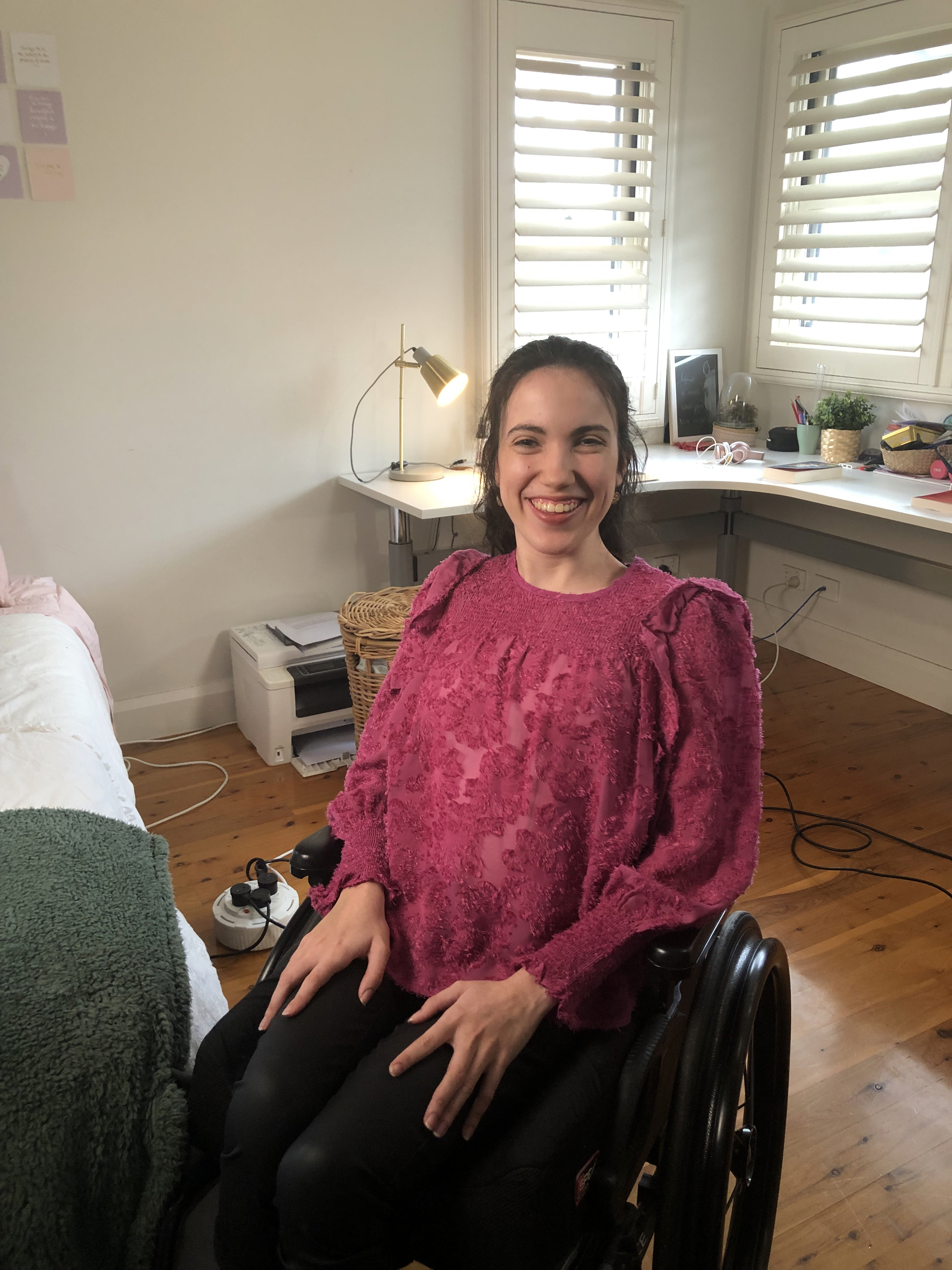 Hannah smiles and is in a room with a long desk which has books, pencils and plants on the surface. She wears a brightly coloured shirt that has embroidery stitched all over it. Hannah is a wheelchair user. 