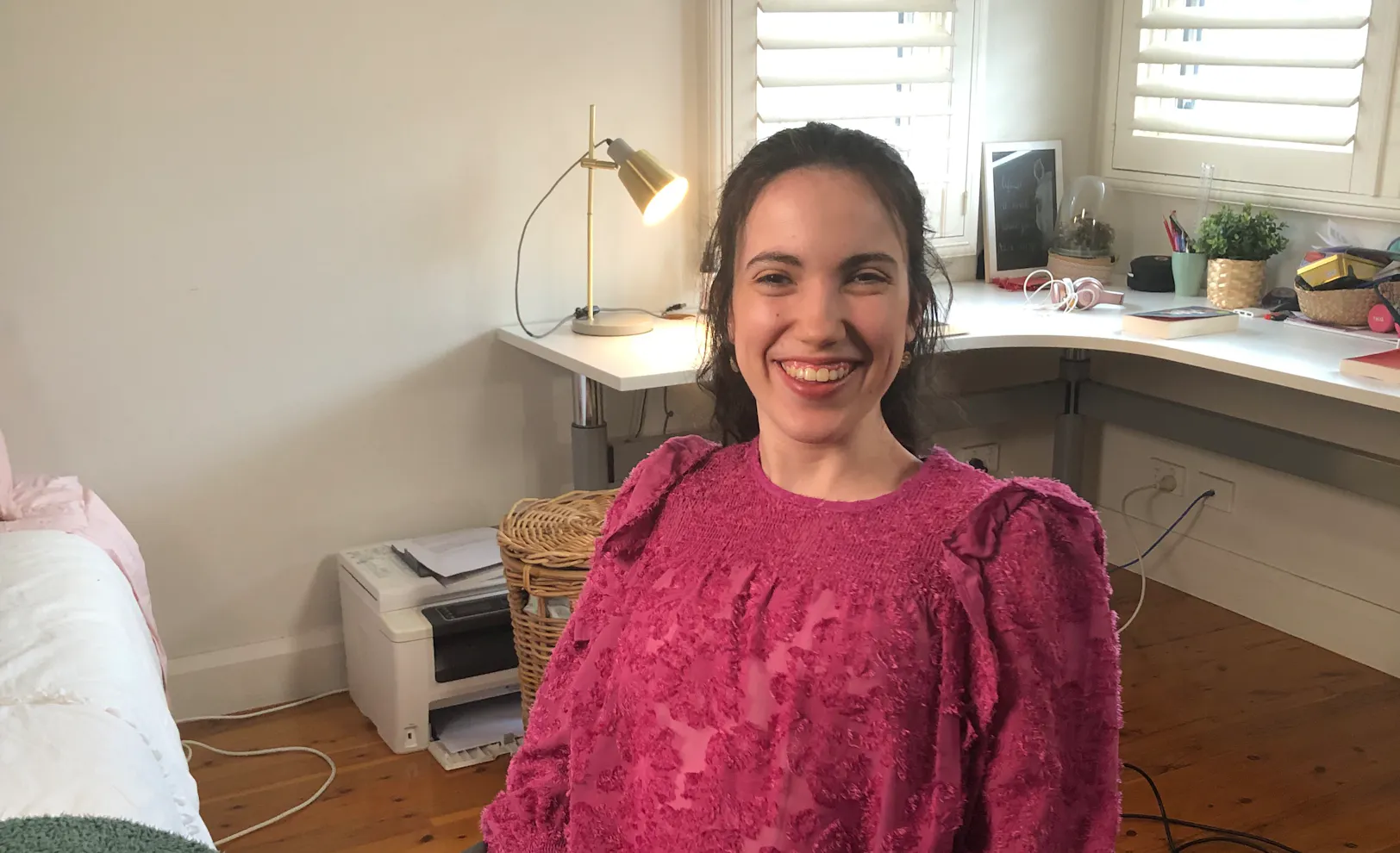 Hannah smiles and is in a room with a long desk which has books, pencils and plants on the surface. She wears a brightly coloured shirt that has embroidery stitched all over it. Hannah is a wheelchair user.