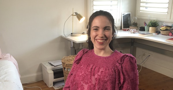 Hannah smiles and is in a room with a long desk which has books, pencils and plants on the surface. She wears a brightly coloured shirt that has embroidery stitched all over it. Hannah is a wheelchair user.
