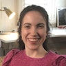 Hannah smiles and is in a room with a long desk which has books, pencils and plants on the surface. She wears a brightly coloured shirt that has embroidery stitched all over it. Hannah is a wheelchair user.