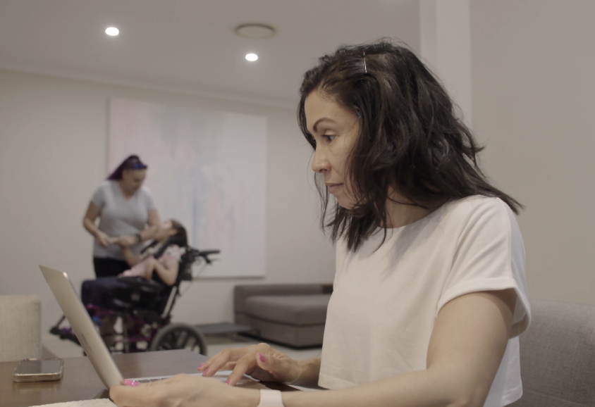Ilse is using her laptop. She has black hair and is wearing a white t-shirt. Jodie is playing with Amara in the background. Jodie is wearing a grey t-shirt and is holding Amara's hand. Amara has brown hair and is a wheelchair user.