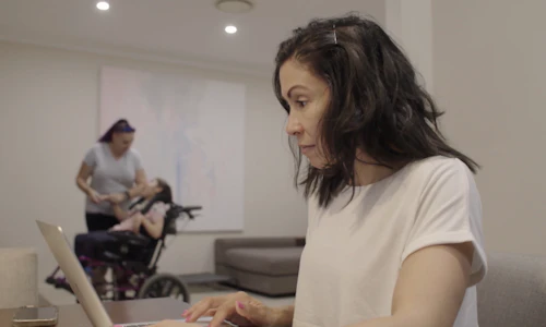 Ilse is using her laptop. She has black hair and is wearing a white t-shirt. Jodie is playing with Amara in the background. Jodie is wearing a grey t-shirt and is holding Amara's hand. Amara has brown hair and is a wheelchair user.