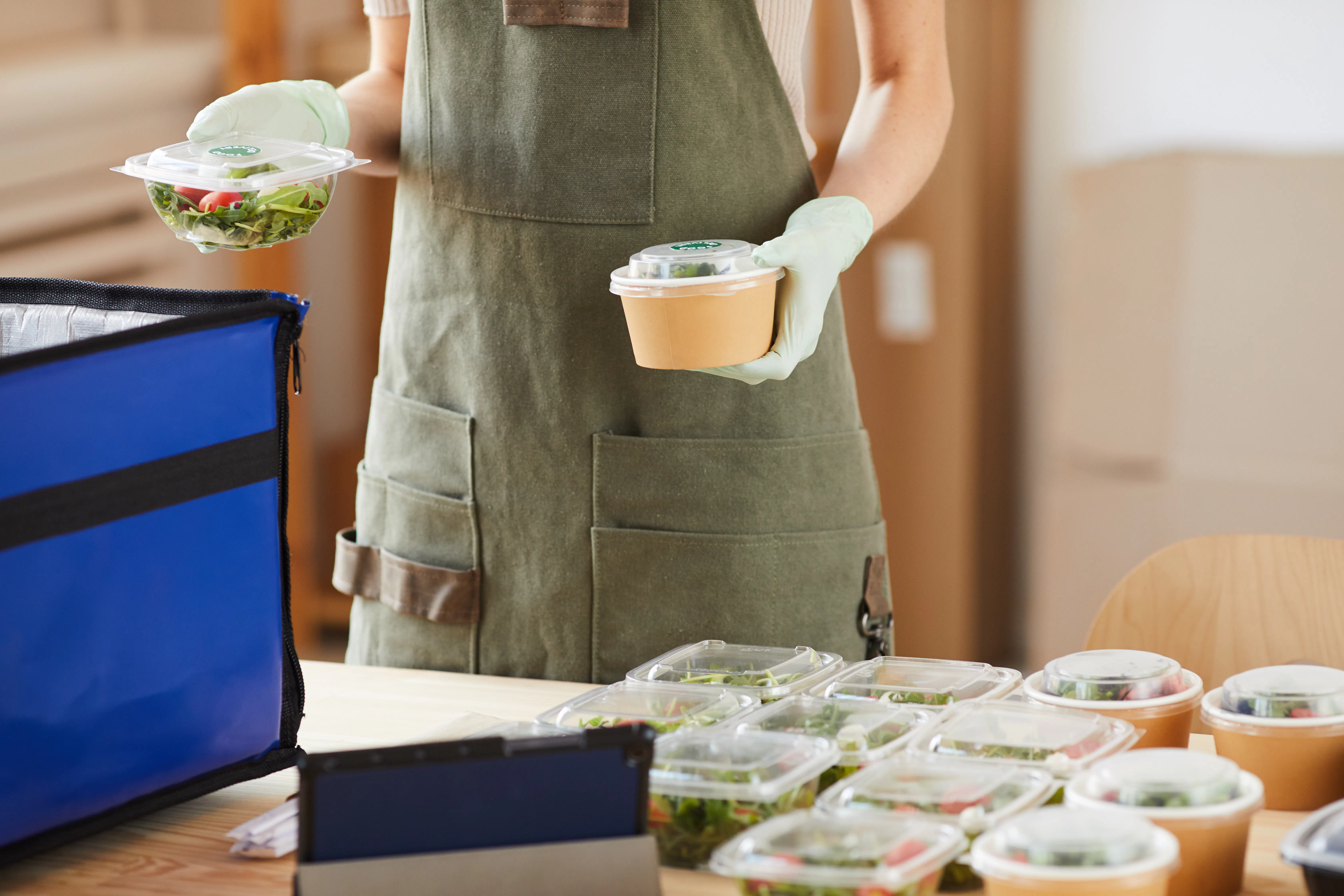 A woman is packing freshly made salads into a blue food delivery cooler bag. She is wearing an olive green apron and disposable gloves.