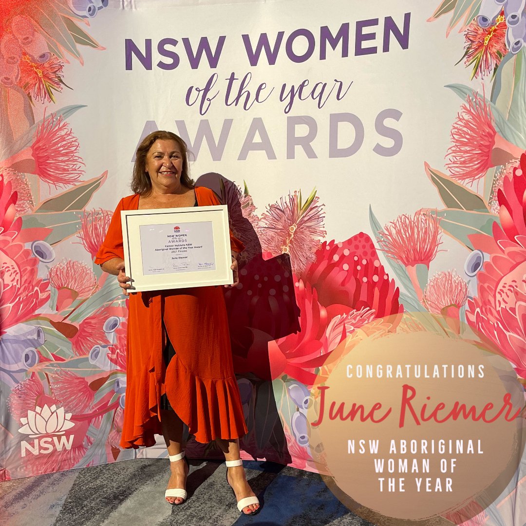 June wears a bright coloured dress and is smiling. She holds a framed award and is in front of a sign  which is covered in botanic flowers and reads 'NSW women of the year awards'. A small circle in the right-hand corner of the photograph reads 'Congratulations June Riemer NSW Aboriginal woman of the year'.