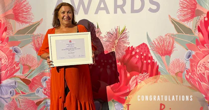 June wears a bright coloured dress and is smiling. She holds a framed award and is in front of a sign which is covered in botanic flowers and reads 'NSW women of the year awards'. A small circle in the right-hand corner of the photograph reads 'Congratulations June Riemer NSW Aboriginal woman of the year'.