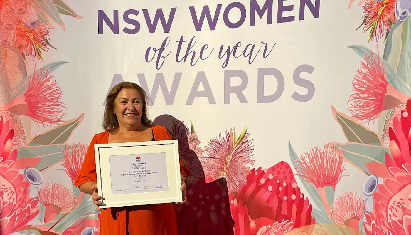 June wears a bright coloured dress and is smiling. She holds a framed award and is in front of a sign which is covered in botanic flowers and reads 'NSW women of the year awards'. A small circle in the right-hand corner of the photograph reads 'Congratulations June Riemer NSW Aboriginal woman of the year'.