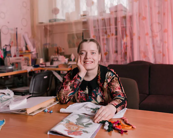 A young woman is sitting at a desk, smiling. One hand is cupping her chin, the other is resting over a piece of embroidery that's almost complete. Pink curtains and a desk covered in craft material is behind her.
