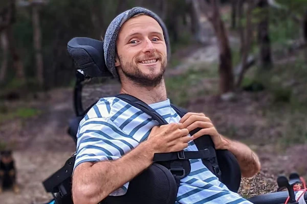 Steve wears a striped shirt, a beanie and is on a dirt track with trees behind him. Steve is smiling at the camera and holding onto his lapel belts that come over his shoulders. Steve is a wheelchair user.
