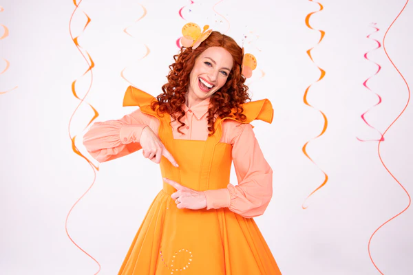 Emma Watkins as 'Emma Memma' wears an orange dress over a peach-coloured blouse. She is signing with Auslan and smiles at the camera.