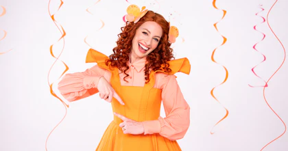 Emma Watkins as 'Emma Memma' wears an orange dress over a peach-coloured blouse. She is signing with Auslan and smiles at the camera.