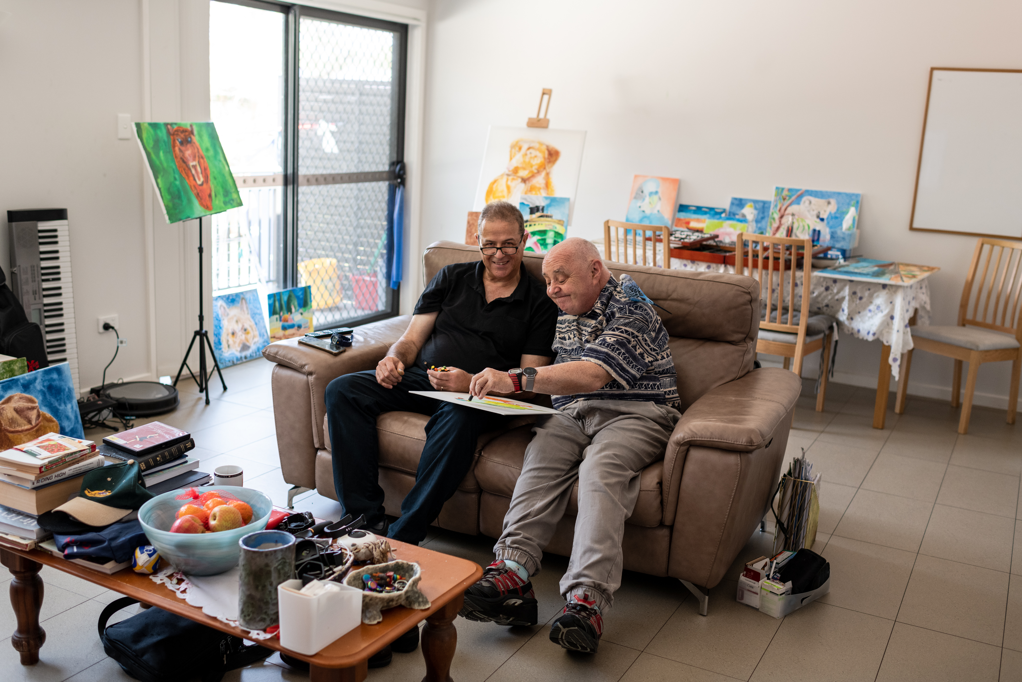 Daniel and Graeme are sitting on a brown couch doing art together with oil pastels. Daniel has short brown hair, wears reading glasses, a black collared shirt and navy trousers. Graeme has short white hair and is wearing two digital watches, a blue and white patterned shirt and grey trousers. 