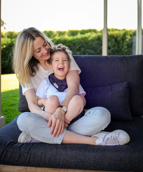 Mel is sitting on a couch with her son Arlo in her lap. They're both laughing.