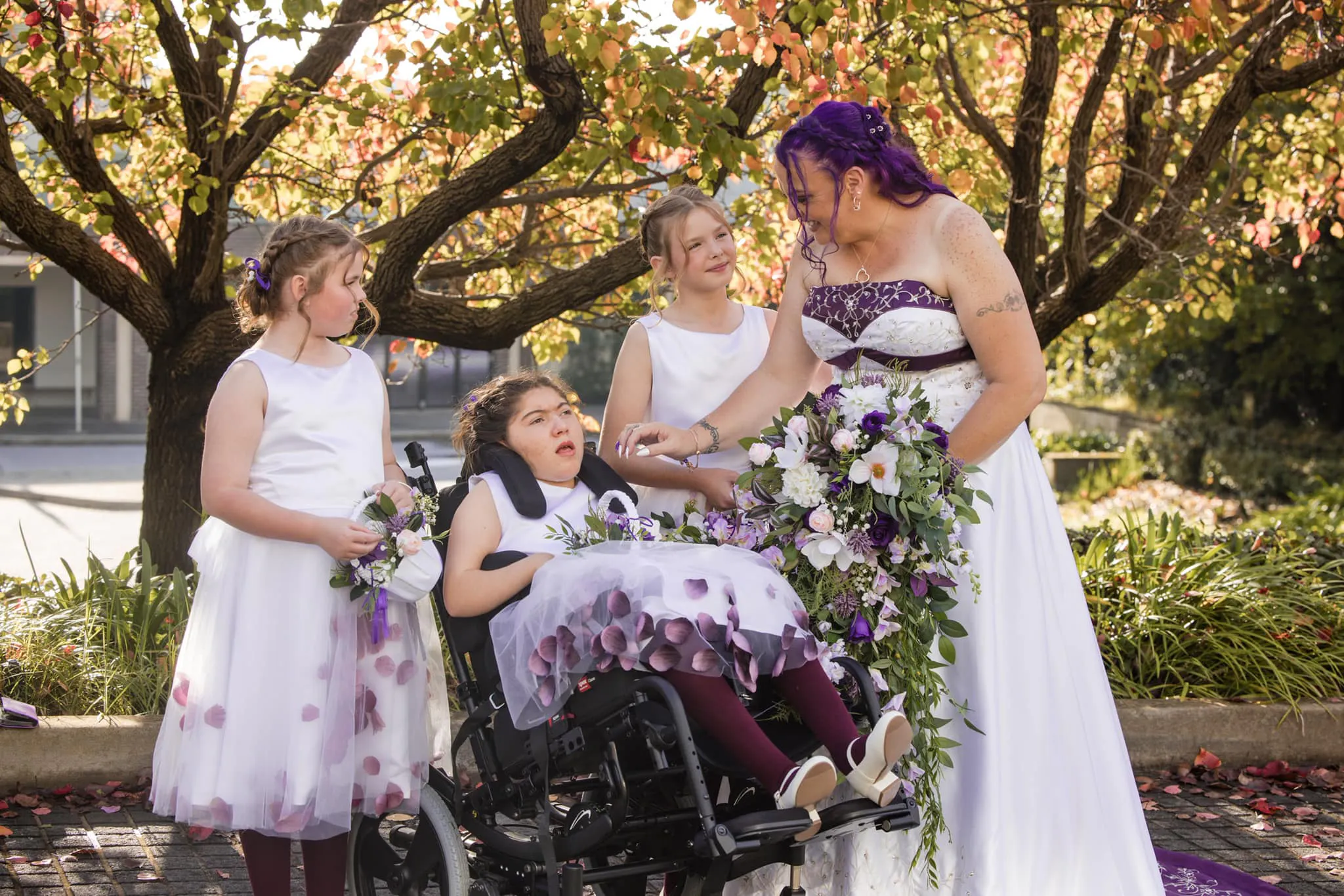 Jodie's three flower girls are all together with Jodie. All are wearing white and purple flower girl dresses and holding white and purple flowers. Jodie is wearing a white and purple wedding dress. Jodie is smiling and reaching out to touch Amara. Amara is a wheelchair user.