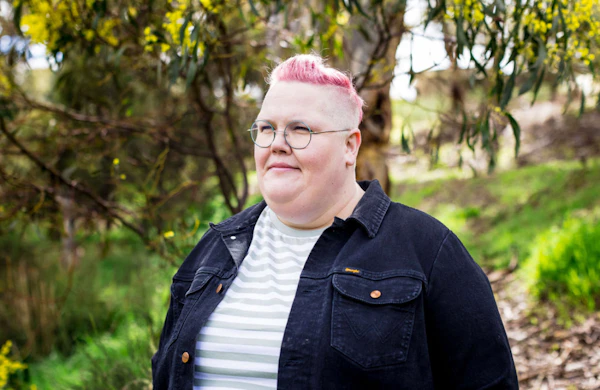 Jasper Peach has short pink hair and wears glasses. They are wearing a grey and white striped top and a navy denim jacket. There is a green eucalyptus tree and bushland.