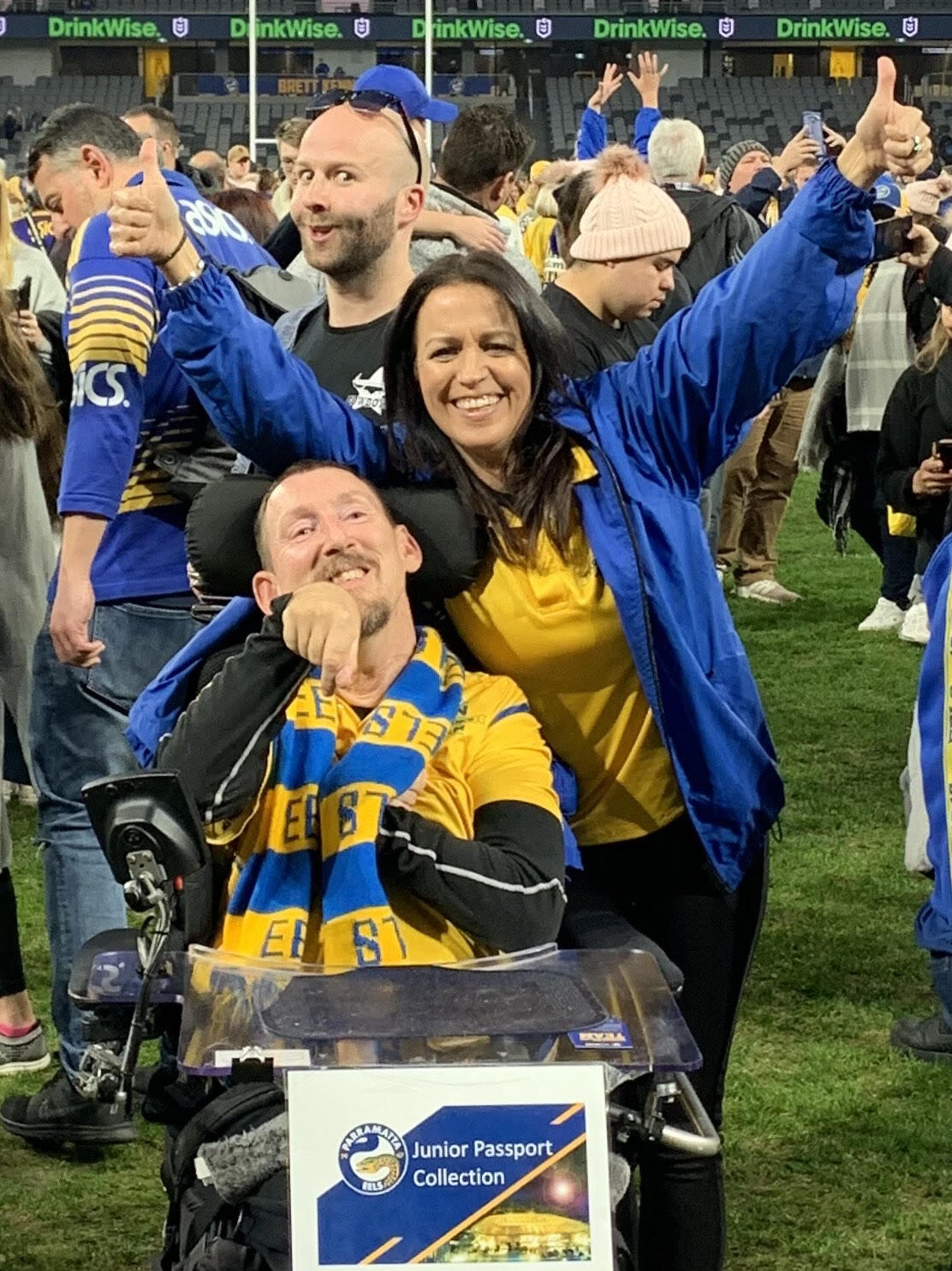Deb and Murray are on the football field wearing all of their Eels merchandise. They are in the middle of cheering.