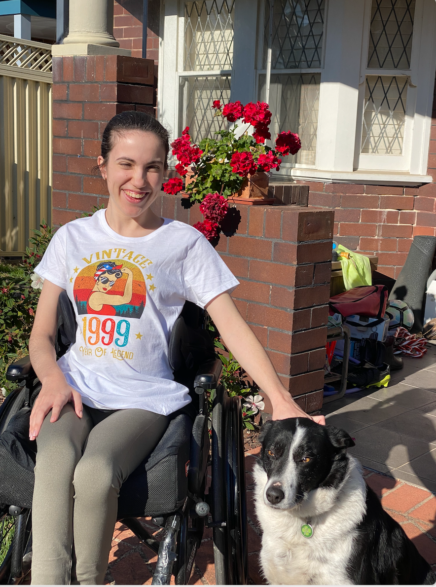 Hannah is wearing a light shirt with a print and dark pants. She is patting a border collie dog sitting beside her. Hannah is a wheelchair user. 