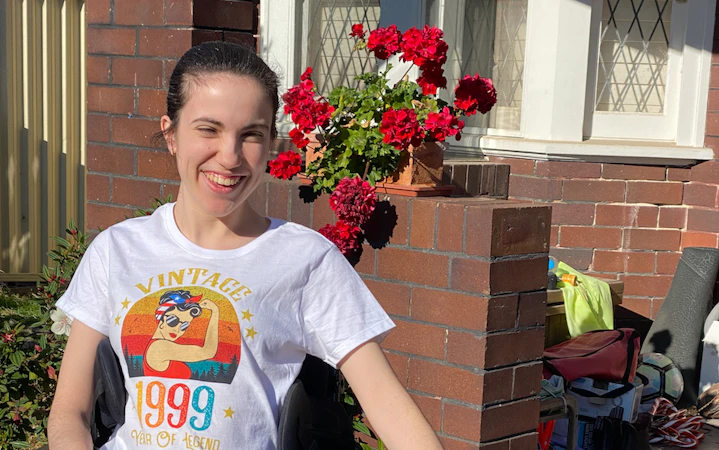 Hannah is wearing a light shirt with a print and dark pants. She is patting a border collie dog sitting beside her. Hannah is a wheelchair user.
