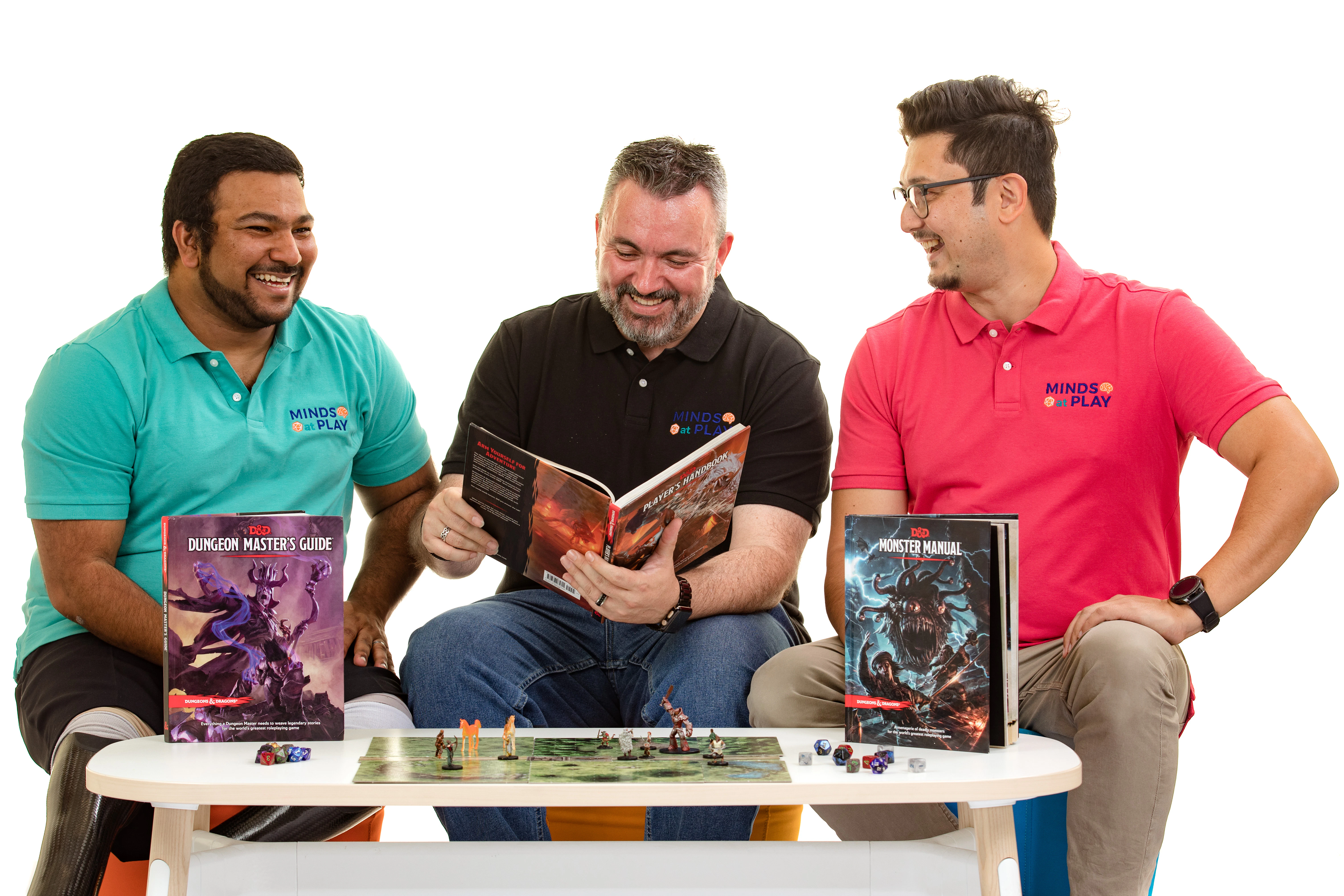 Three men sit around a coffee table, smiling. They each wear a collared shirt with their organisation name 'Minds at Play' embroidered on the top, right side of the shirt. A board game is set up on the table. One of the men is holding and reading a games book.