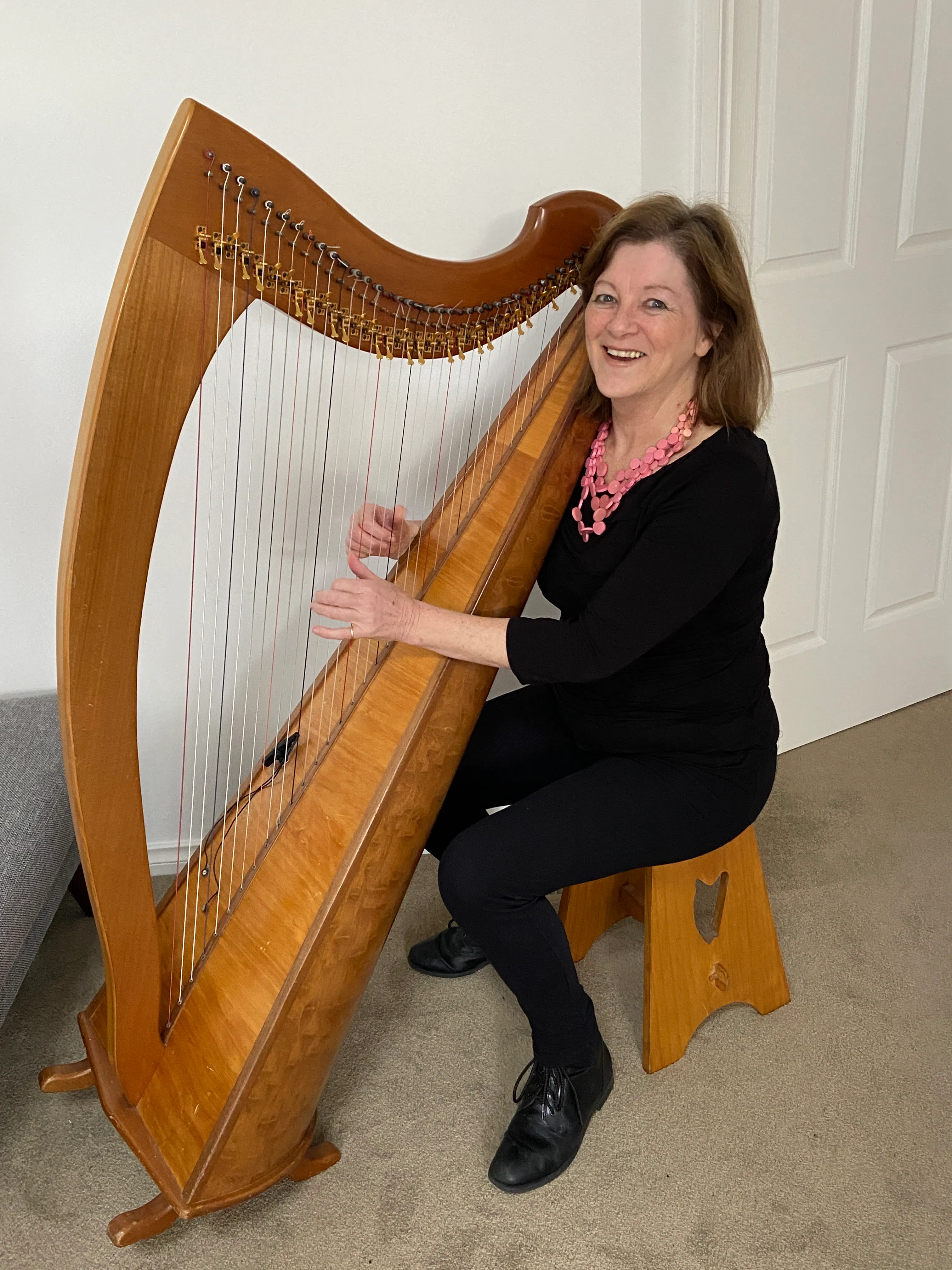 A woman is wearing a black long sleeve dress and a pink beaded necklace. She is strumming a large harp that has a wooden frame. 