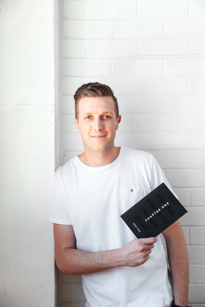 Daniel Flynn is leaning against a white wall holding a black book. Daniel has short hair and is wearing a white t-shirt.