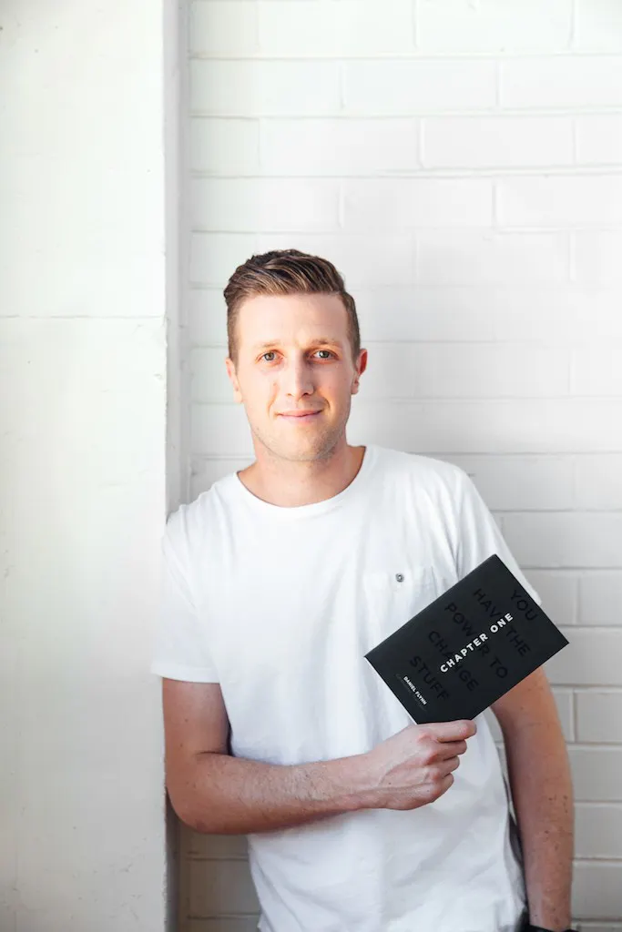 Daniel Flynn is leaning against a white wall holding a black book. Daniel has short hair and is wearing a white t-shirt.