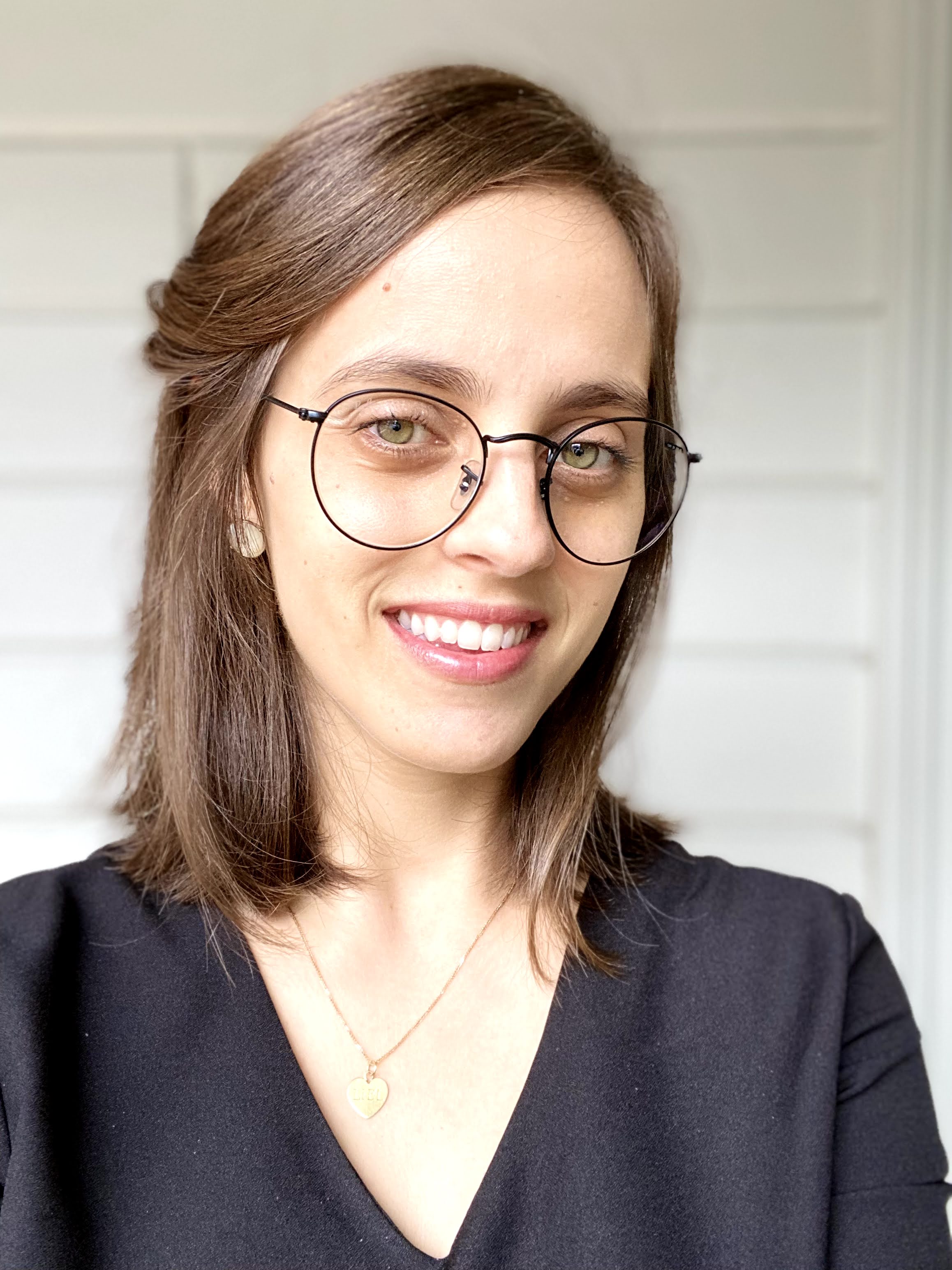 Liel Bridgford has shoulder-length brown hair and green eyes. She wears glasses, a golden heart locket and a dark top. 