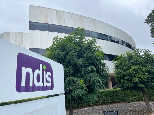 A grey building surrounded by a green hedge and trees is pictured behind a sign featuring the NDIS logo. 