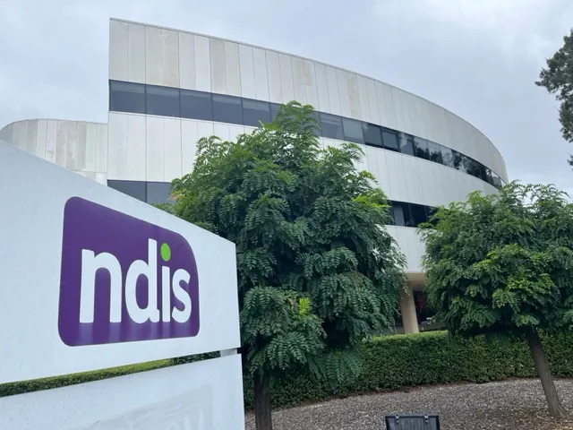 A grey building surrounded by a green hedge and trees is pictured behind a sign featuring the NDIS logo. 